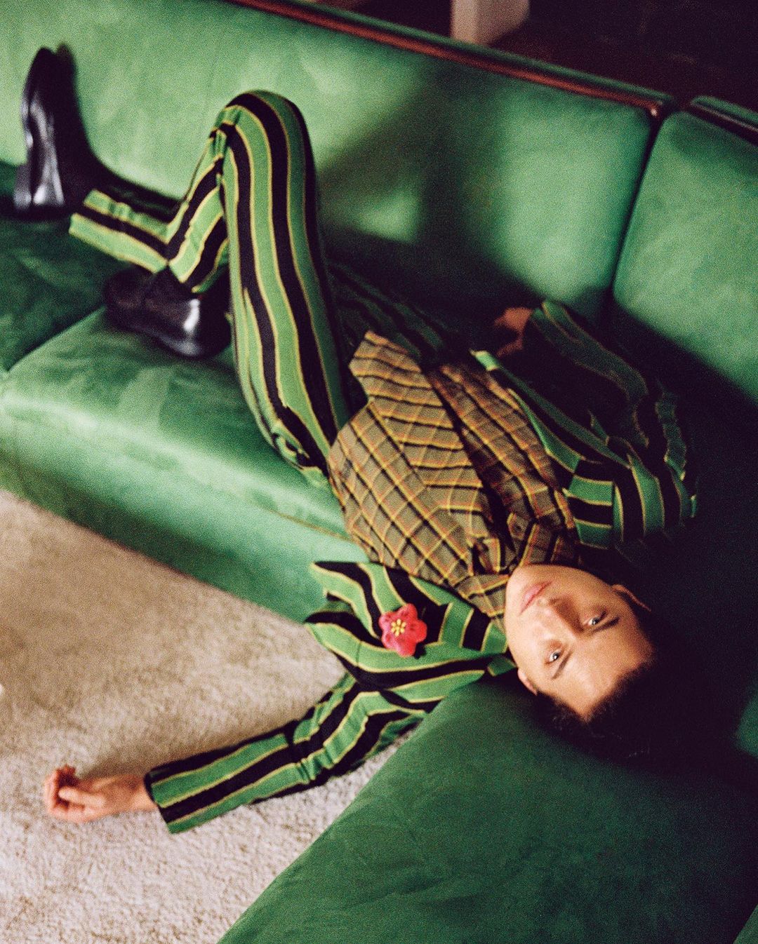 Adult male laying on a green velvet couch wearing a striped suit jacket and pants and black shoes in an indoor setting.