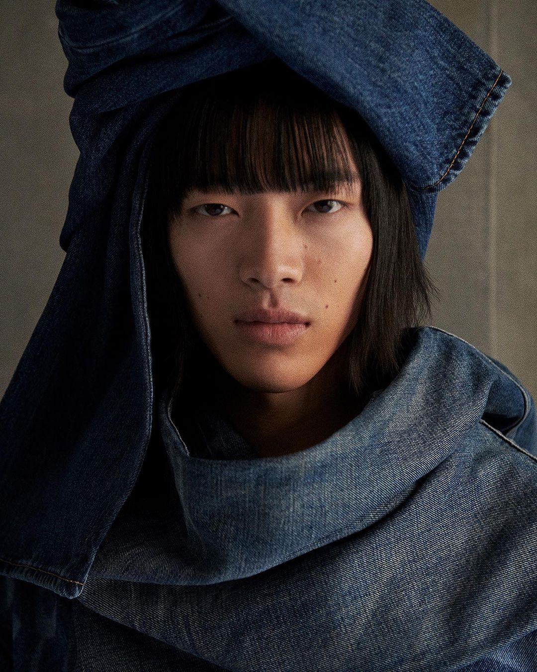 Close-up portrait of a young Asian person with dark hair and bangs, wearing a denim garment draped around the head and neck against a neutral background.