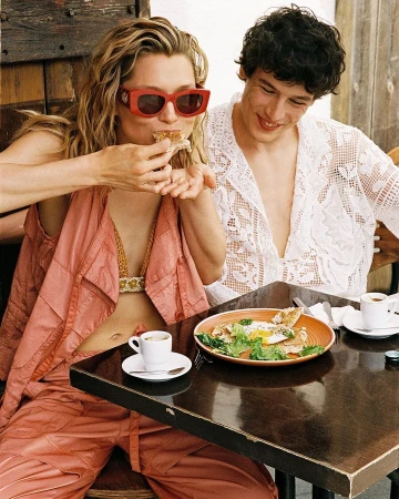 Young couple enjoying a meal at an outdoor cafe, woman eating sandwich, man smiling, sunny day, table with food and coffee.