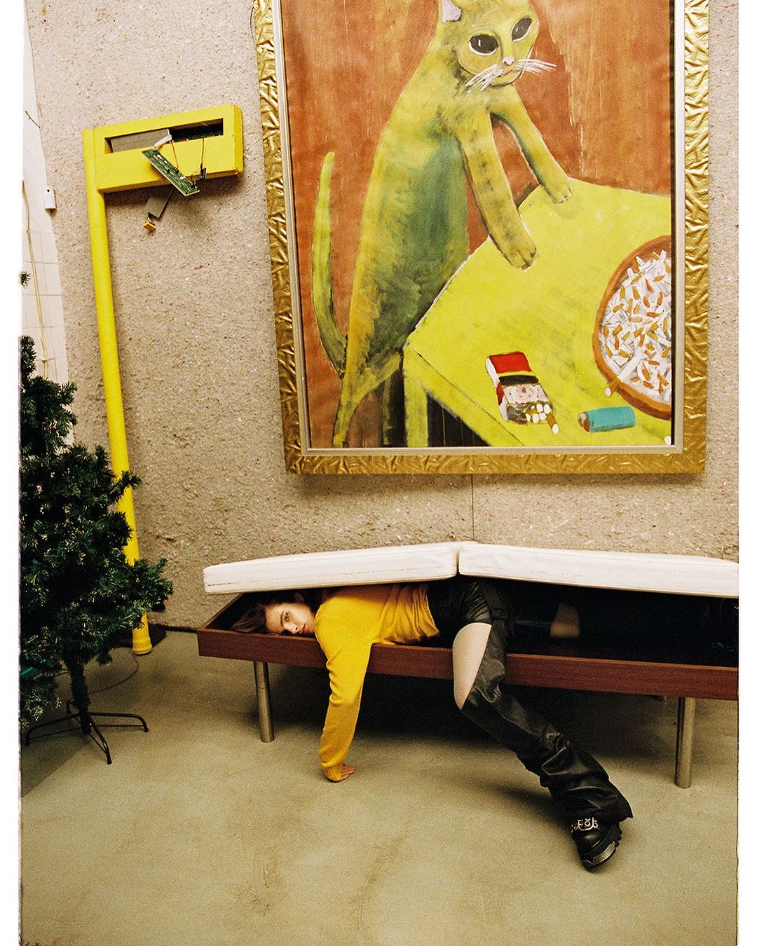 Woman in a yellow shirt and black leather boots poses under a bench with a cat painting and yellow decoration on the wall.