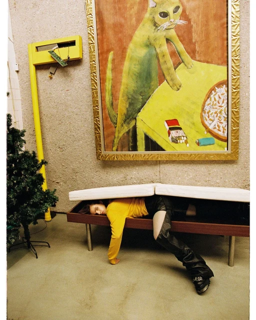 Woman in a yellow shirt and black leather boots poses under a bench with a cat painting and yellow decoration on the wall.