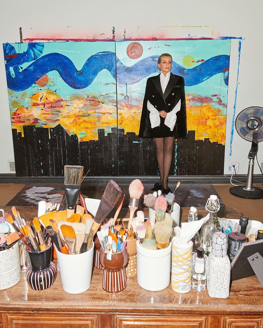Blonde woman poses in a black and white cape blazer in front of an abstract painting in an artist's studio filled with paintbrushes.