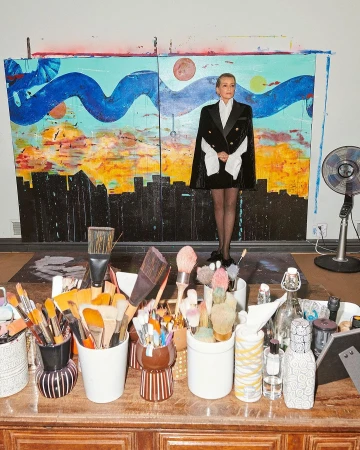 Blonde woman poses in a black and white cape blazer in front of an abstract painting in an artist's studio filled with paintbrushes.