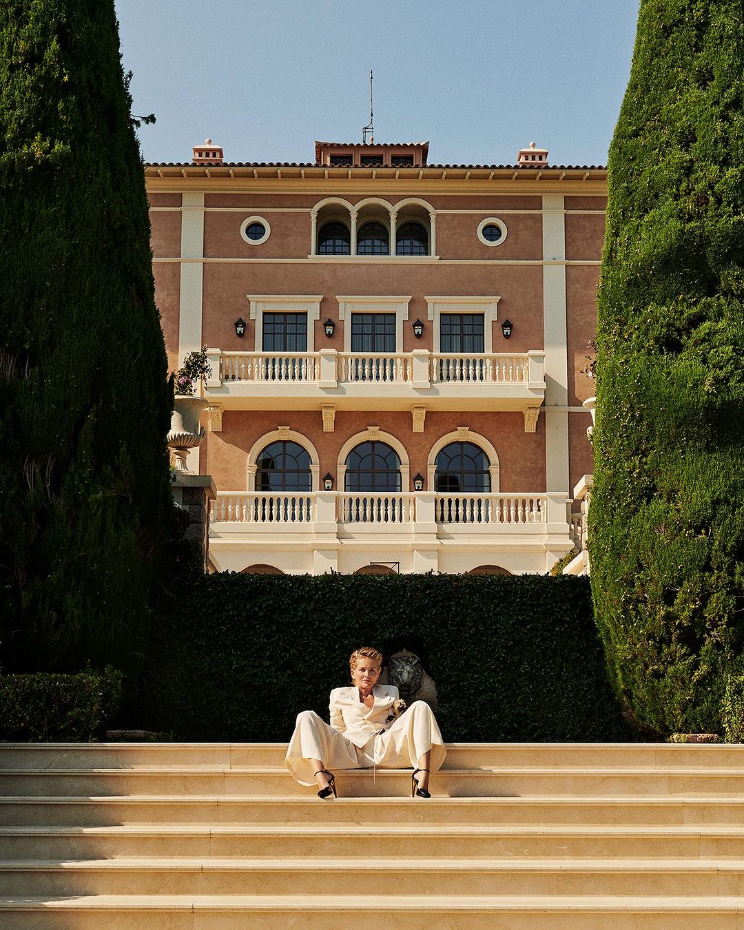 Woman in white suit sits on stairs in front of a large building framed by tall trees against a blue sky in a wide outdoor shot.