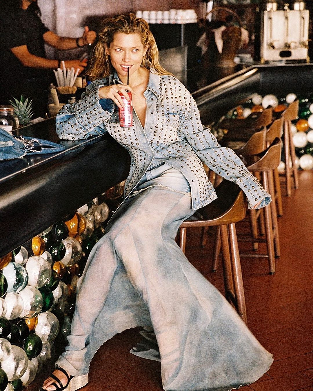 Woman in embellished denim jacket and sheer skirt seated at bar, sipping drink with straw. Interior bar setting with unique decorations.