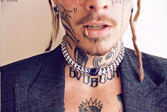 Close-up portrait of a tattooed adult male with dreadlocks, wearing a silver chain necklace and blazer against a neutral background.