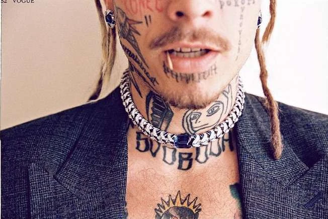 Close-up portrait of a tattooed adult male with dreadlocks, wearing a silver chain necklace and blazer against a neutral background.