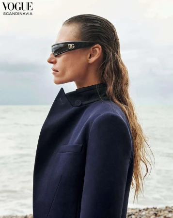 Model wearing dark sunglasses and navy blazer posing outdoors near a sandy beach with overcast sky background