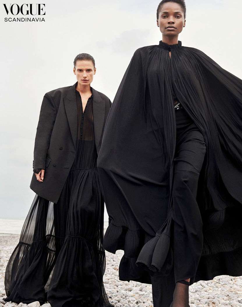 Two women in black dresses pose on a rocky shore for Vogue Scandinavia, showcasing flowing fabric and dark fashion against a neutral sky.