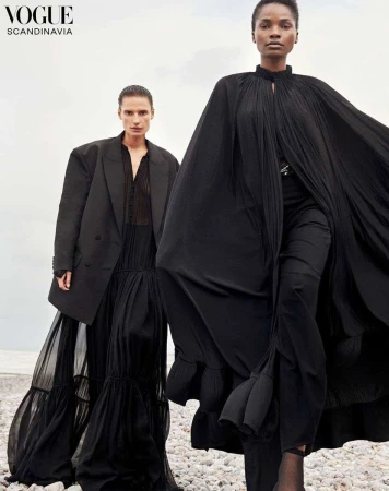 Two women in black dresses pose on a rocky shore for Vogue Scandinavia, showcasing flowing fabric and dark fashion against a neutral sky.