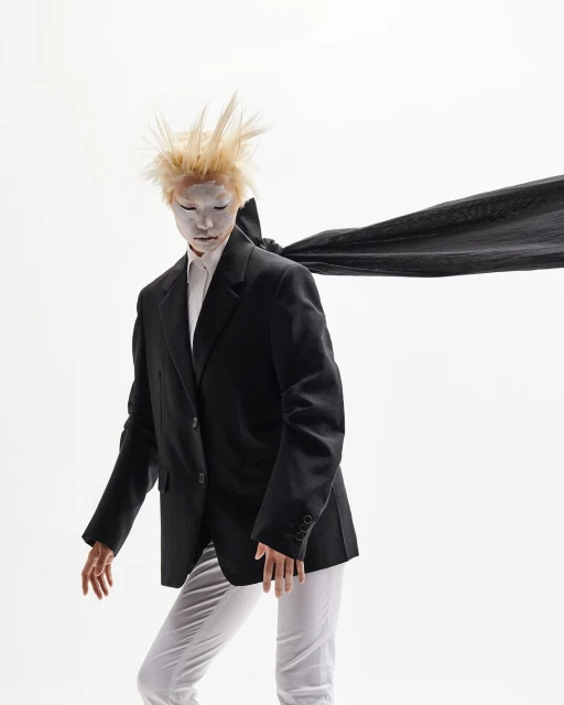 Fashion shot of model wearing a black suit jacket with white makeup and spiky blonde hair, posing against a white background.