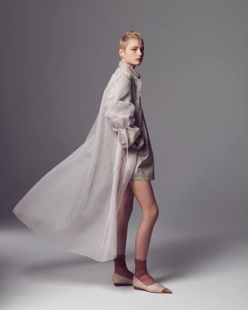 Model with short blonde hair wearing a sheer flowing coat and flats against a grey seamless background