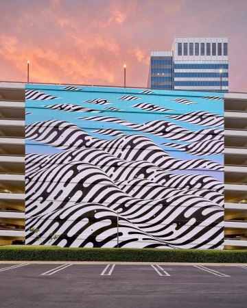 Large-scale optical illusion mural adorns a building, featuring wavy black and white stripes against a pink and blue sky backdrop.