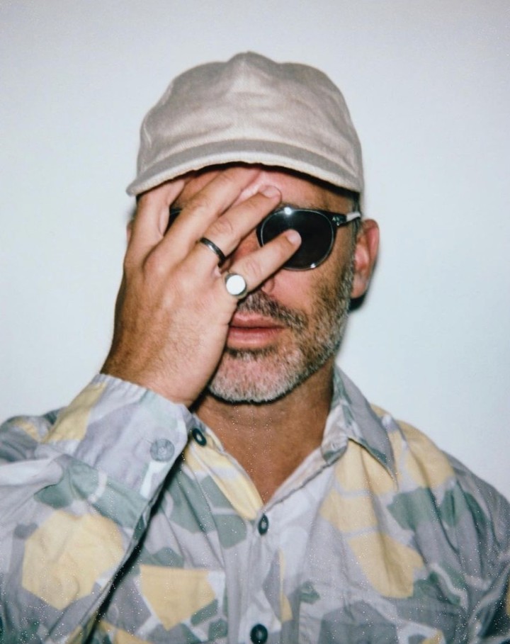 Man wearing a patterned shirt, cap, and sunglasses with his hand partially covering his face against a white wall.