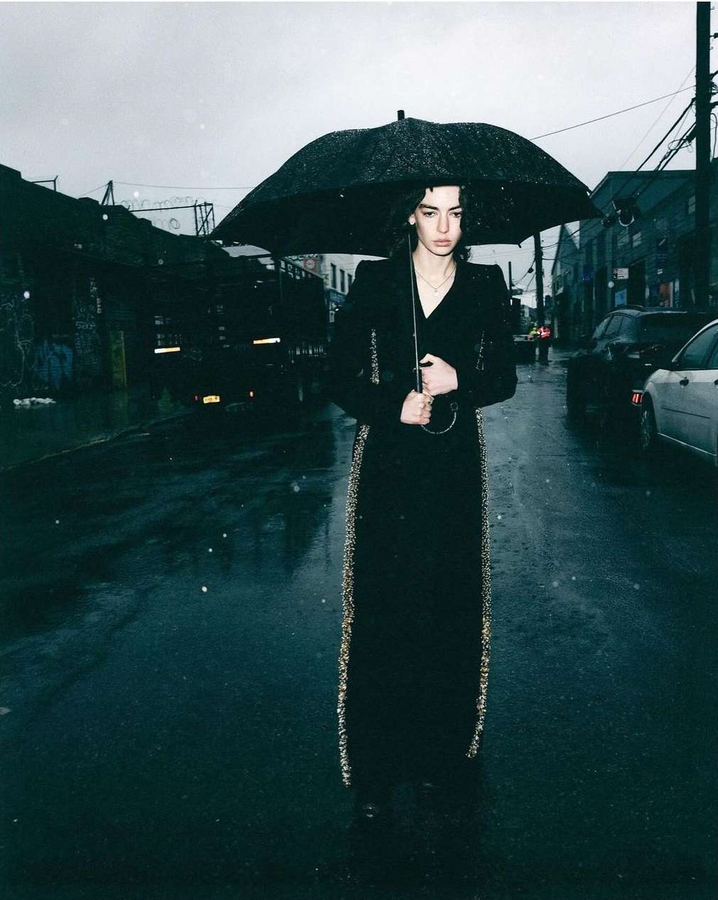 A person in a long black dress and coat stands on a wet street under a large black umbrella in rainy conditions.