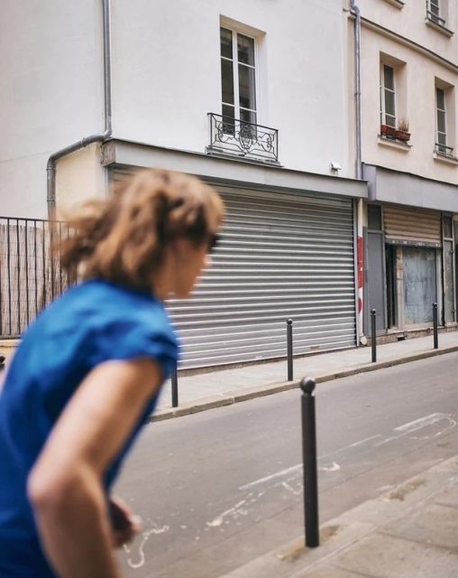 Blurred action shot of a woman running on a city street in front of white buildings and closed storefronts with security shutters.