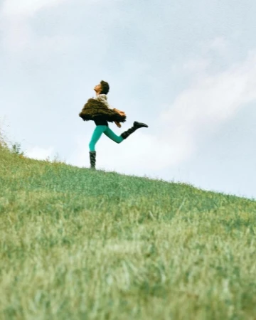 A woman in a green bodysuit and furry vest runs uphill through a grassy field under a partly cloudy sky, captured in a dynamic pose.