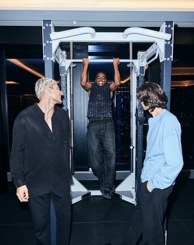Three men in an indoor gym setting: one doing pull-ups, and two others standing by. Dark ambiance with a city view visible outside.