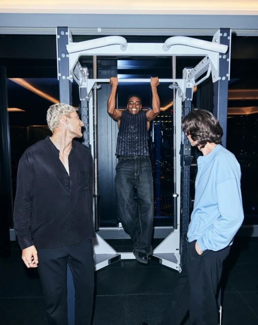 Three men in an indoor gym setting: one doing pull-ups, and two others standing by. Dark ambiance with a city view visible outside.