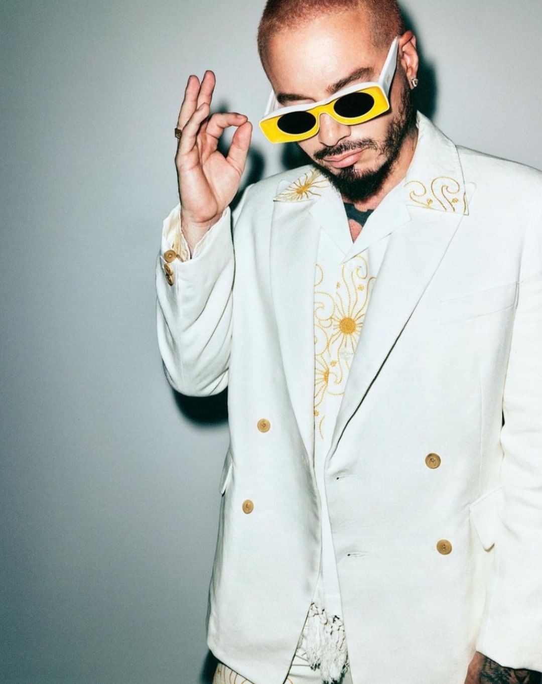 Adult male wearing a white suit and yellow-framed sunglasses gestures with his hand in a studio setting against a grey backdrop.