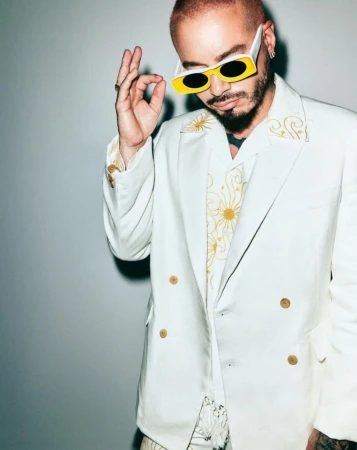 Adult male wearing a white suit and yellow-framed sunglasses gestures with his hand in a studio setting against a grey backdrop.