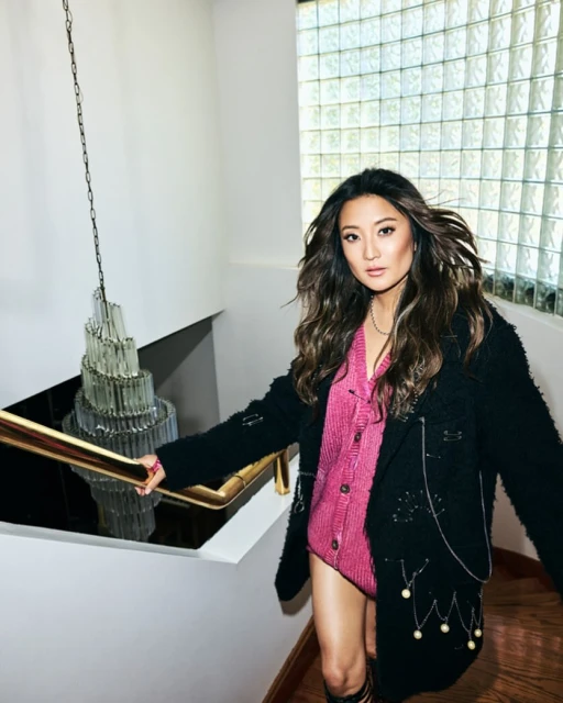 Asian woman poses on stairs in a pink bodysuit, black jacket, and tall socks. Modern interior with crystal chandelier and light from window.