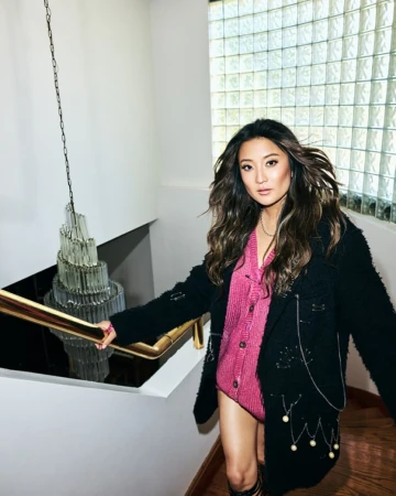 Asian woman poses on stairs in a pink bodysuit, black jacket, and tall socks. Modern interior with crystal chandelier and light from window.