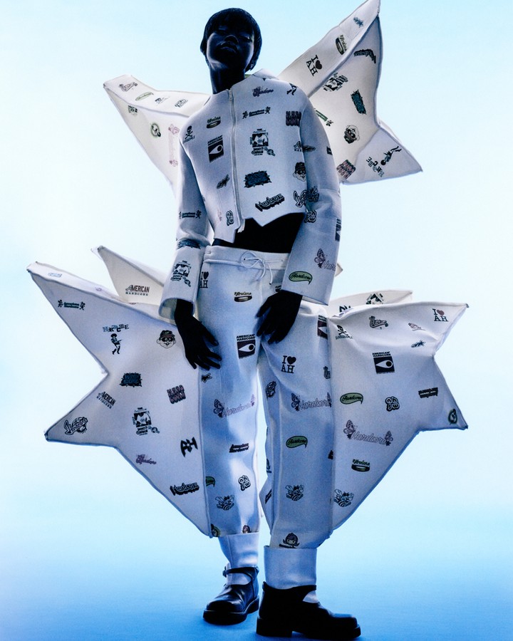A person is standing in a studio with an avant-garde outfit and black boots against a light blue background in a fashion photography style.