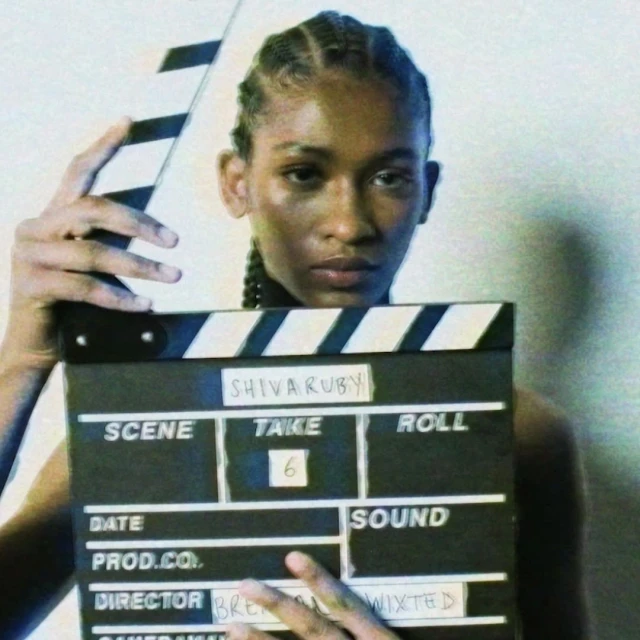 Adult woman holds a film slate indoors, which features production details. The film slate divides the composition in half vertically.