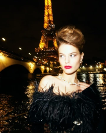 Woman with red lipstick in feathered dress poses in Paris with Eiffel Tower and the Seine River at night with bridge lights.