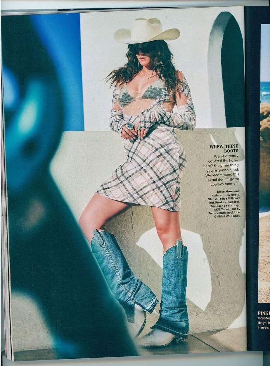 A woman poses in cowboy boots and a hat, wearing a plaid dress and sheer top, set against a backdrop of white walls and a beach.