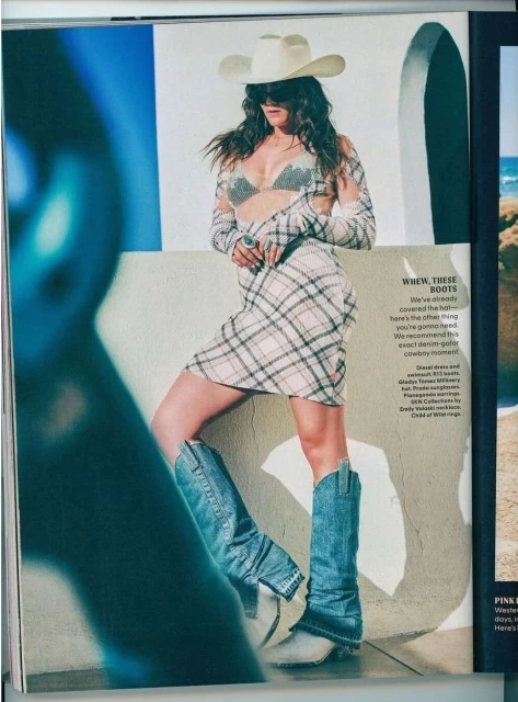 A woman poses in cowboy boots and a hat, wearing a plaid dress and sheer top, set against a backdrop of white walls and a beach.