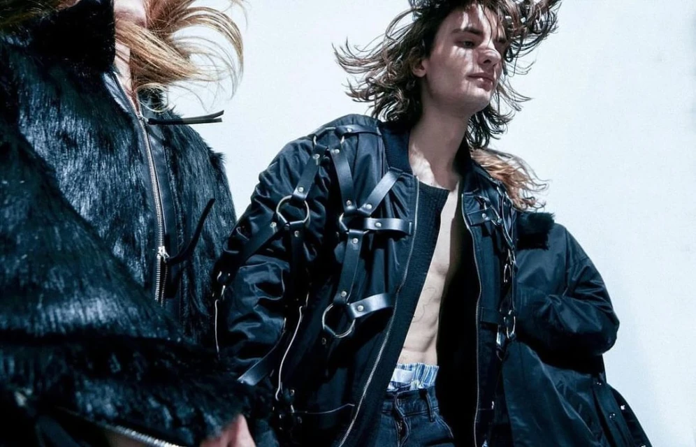 Two fashion models pose indoors in a photo studio wearing black jackets with leather straps, complemented by a light backdrop.