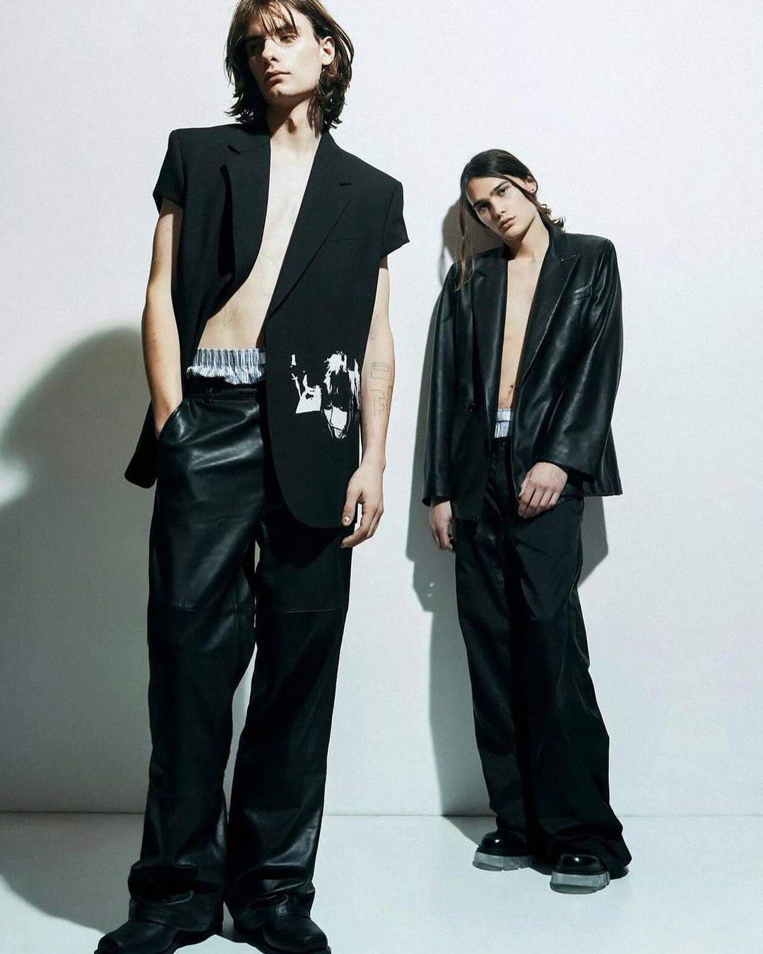 Two slender male models in black blazers and loose pants pose indoors against a white wall for a fashion editorial shoot.