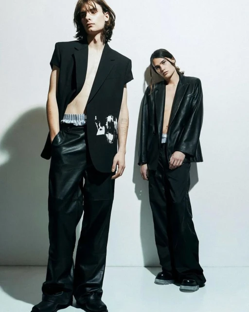 Two slender male models in black blazers and loose pants pose indoors against a white wall for a fashion editorial shoot.