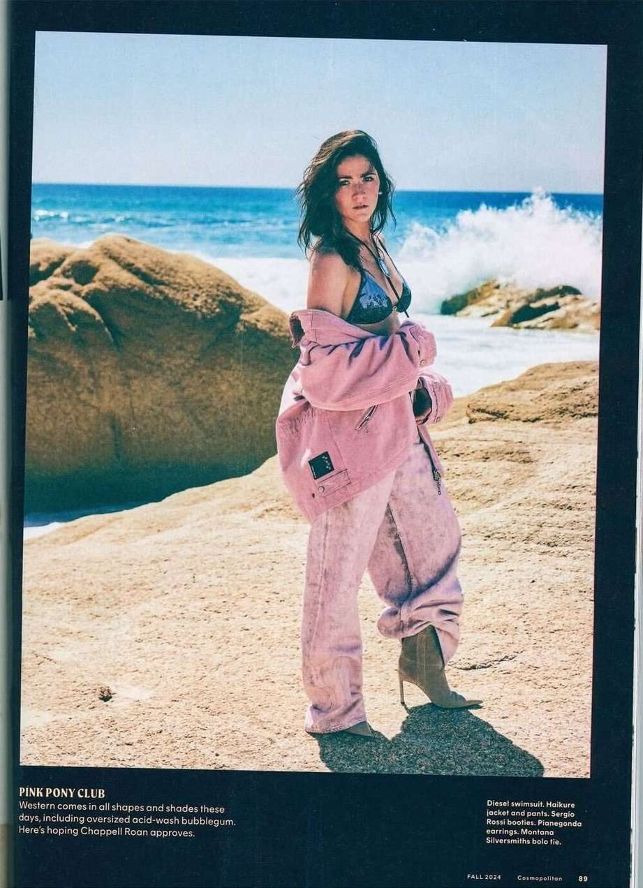 A fashionable woman in a bikini top, pink jacket, and light pink pants poses on a beach with ocean waves and rocks in the background.