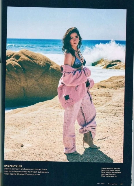 A fashionable woman in a bikini top, pink jacket, and light pink pants poses on a beach with ocean waves and rocks in the background.