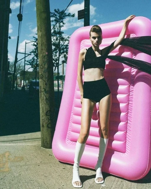 An adult fashion model poses outdoors in a polka dot top and briefs with an inflatable pink pool raft on a city street.