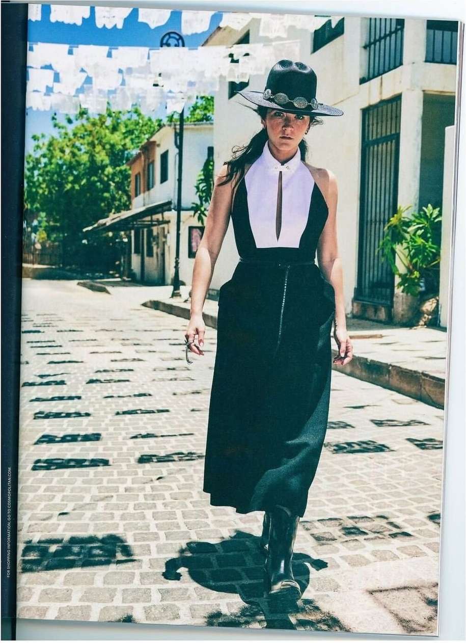 A stylish woman in a black-and-white dress and black hat walks down a cobblestone street lined with white buildings and festive decorations.