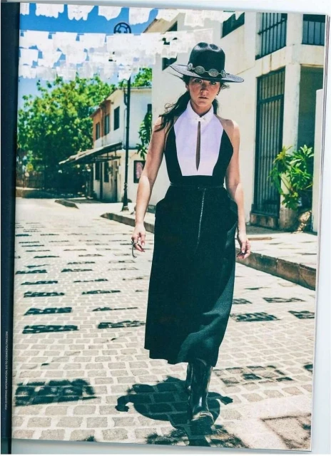 A stylish woman in a black-and-white dress and black hat walks down a cobblestone street lined with white buildings and festive decorations.