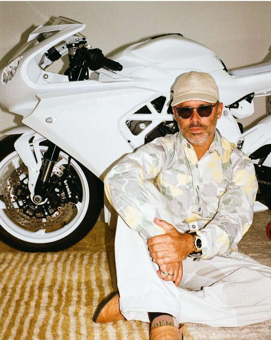 An adult man sits in front of a white motorcycle wearing a patterned button-down shirt, white pants, sunglasses, and a beige cap.