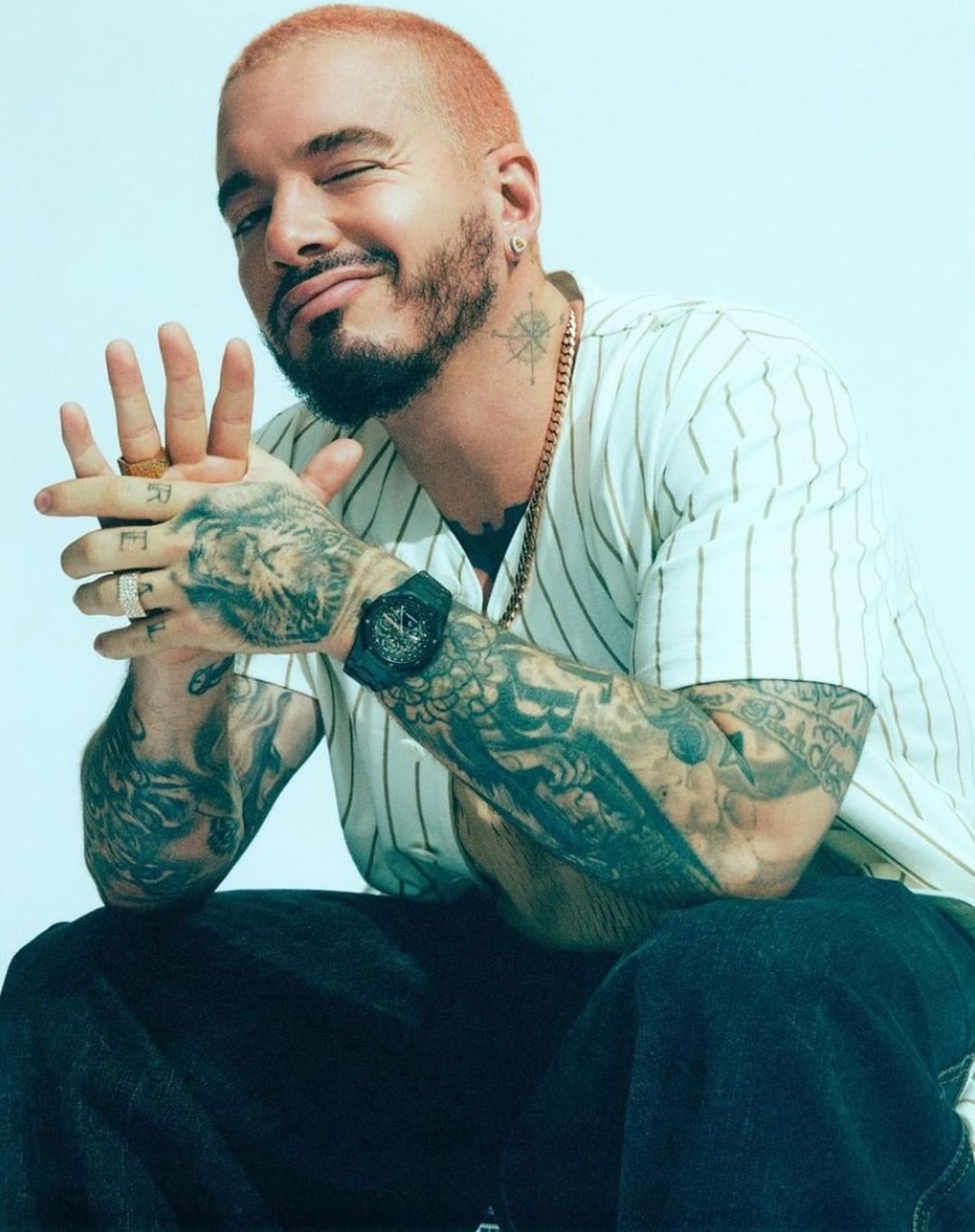 Portrait of an adult male with short red hair, tattoos on his arms, a black wristwatch, gold chains, and blue jeans in a studio.