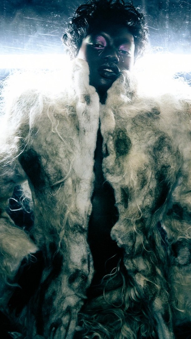 Close-up of a fashion model with striking makeup wearing a fuzzy, textured coat, set against a backdrop with a bright, blurry light.