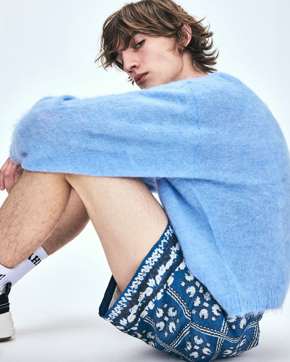 Young male model seated wearing a fuzzy light blue sweater and patterned blue shorts against a white backdrop.