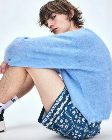 Young male model seated wearing a fuzzy light blue sweater and patterned blue shorts against a white backdrop.