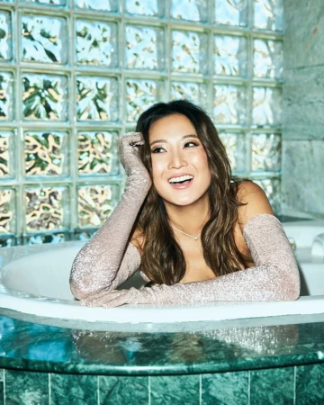Smiling Asian woman in bathtub with sparkly long gloves, set against a backdrop of clear glass blocks with leafy patterns.