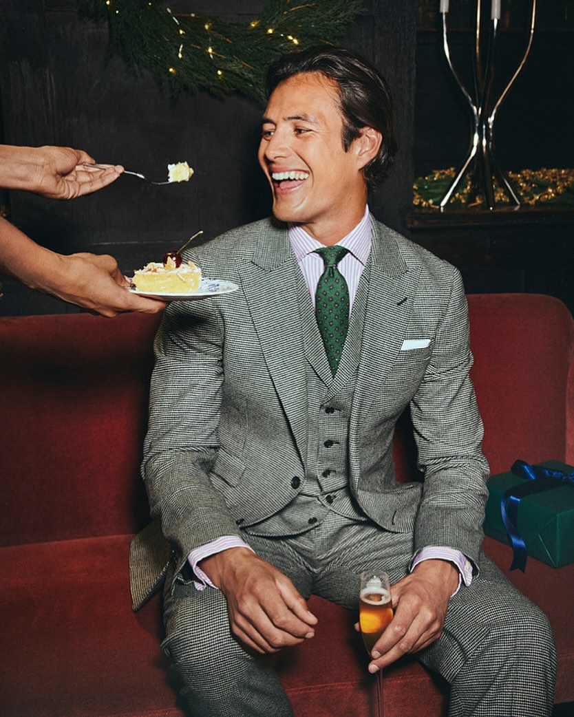 Smiling man in a suit is offered a slice of cake. He is holding a glass of beer while seated on a red couch indoors.