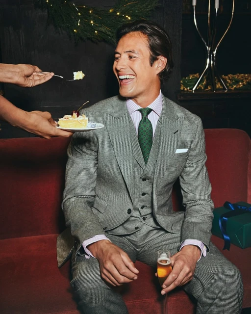 Smiling man in a suit is offered a slice of cake. He is holding a glass of beer while seated on a red couch indoors.