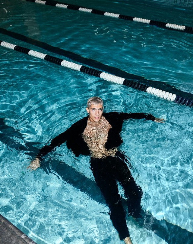 Person floats in a swimming pool wearing a black jacket and pants, water ripples on body, pool lane dividers are in the background.