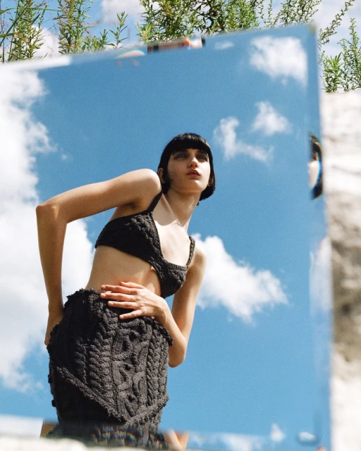 A young woman poses in matching knit top and skirt in front of a mirror reflecting the blue sky with green foliage above.